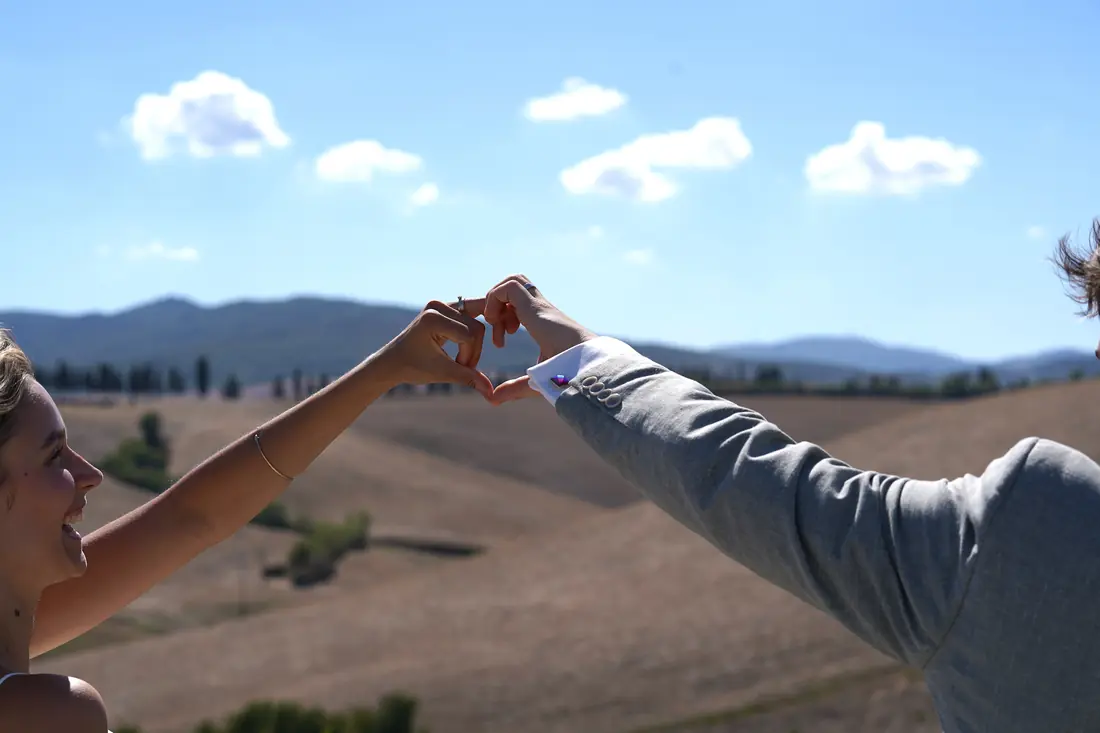 The couple making a heart with their hands over the Tuscan hills